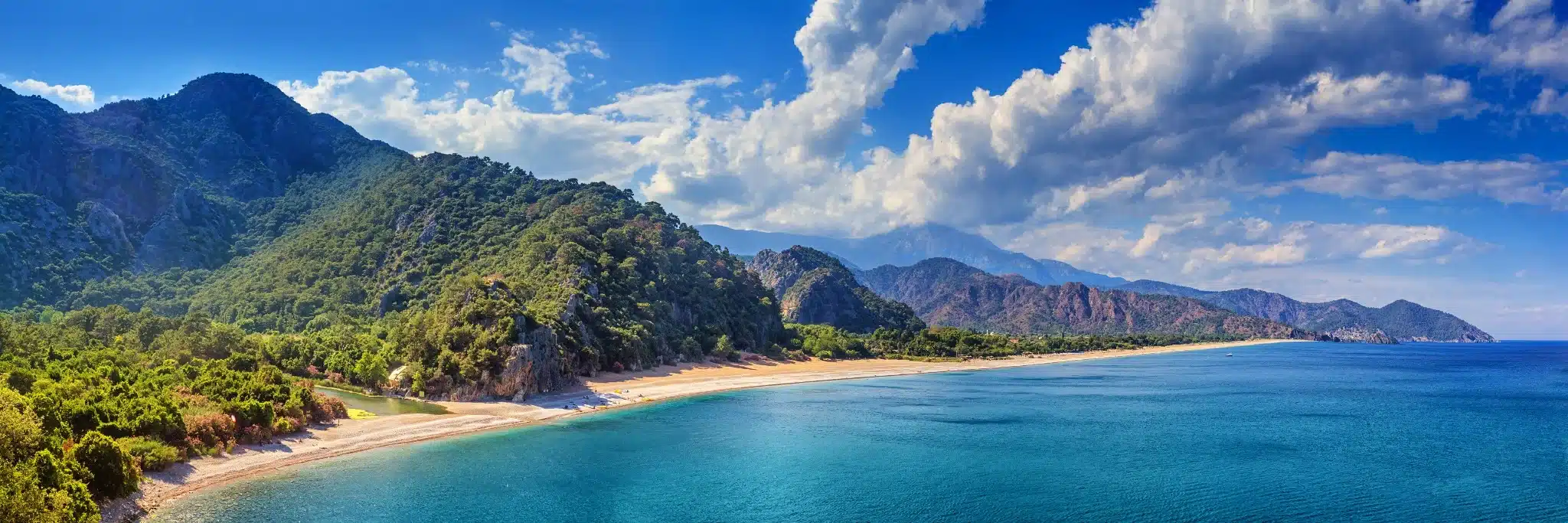 Panoramisch uitzicht over het strand van Olympos in Kemer, Antalya, met turquoise zee, groene bergen en een uitgestrekte kustlijn onder een blauwe hemel met wolken.