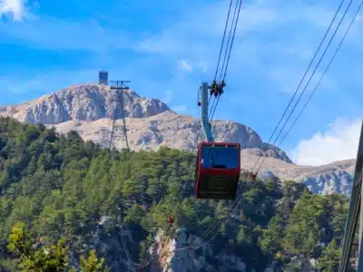 Olympos Teleferik – Tahtalı Kabelbaan Rode gondel van de Olympos Teleferik (Tahtalı Kabelbaan) die omhoog gaat naar de top van de Tahtalı Dağı met dennenbossen en rotsachtige bergen op de achtergrond.