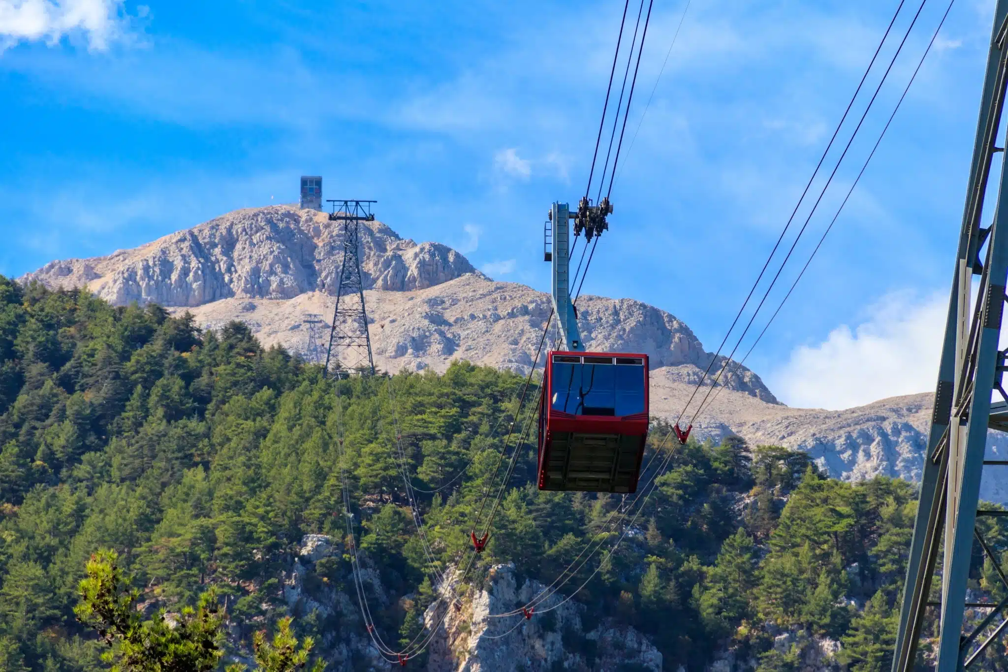 Rode gondel van de Olympos Teleferik (Tahtalı Kabelbaan) die omhoog gaat naar de top van de Tahtalı Dağı met dennenbossen en rotsachtige bergen op de achtergrond.